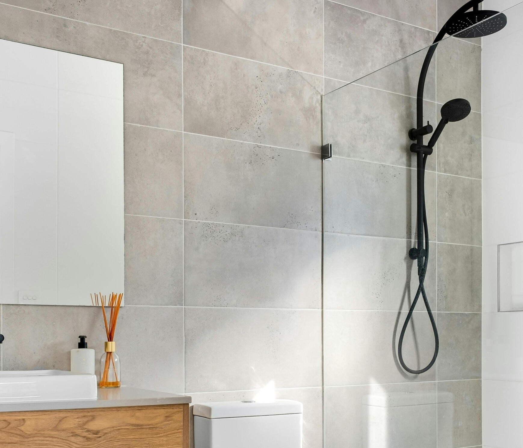 Modern bathroom with light‑grey tiles, black shower fixtures, wooden vanity, and minimalist design A clean, contemporary bathroom featuring large light‑grey tiles, a black rainfall showerhead, a wooden vanity with a white countertop, and a modern toilet. This style reflects the premium yet affordable renovation work offered by Sydney Budget Kitchens.